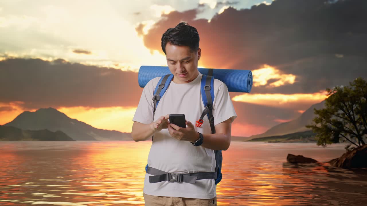 Asian Male Hiker With Mountaineering Backpack Enjoy Using Smartphone At A Lake