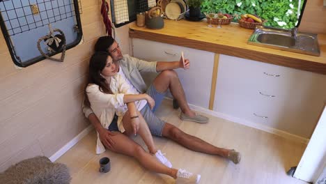 beautiful couple sitting on the floor in the stylish kitchen in van and doing selfie using smartphone, posing . enjoying togetherness, shared holidays, traveling by wheels house. modern interior. high angle footage