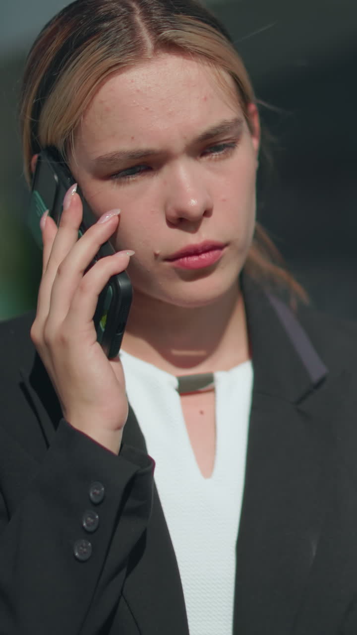 Thoughtful woman in formal attire walks during phone call, focused expression under sunlight, with blur background featuring building with glass facade and reflective surfaces in urban setting