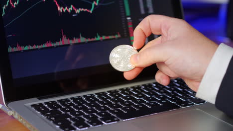 el hombre de negocios voltea la moneda de cardarno en sus dedos mientras mira los gráficos de cambio y los precios en una computadora portátil