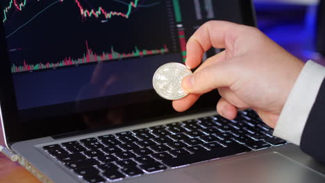 businessman flips over cardarno coin in his fingers whilst looking at the exchange charts and prices on a laptop