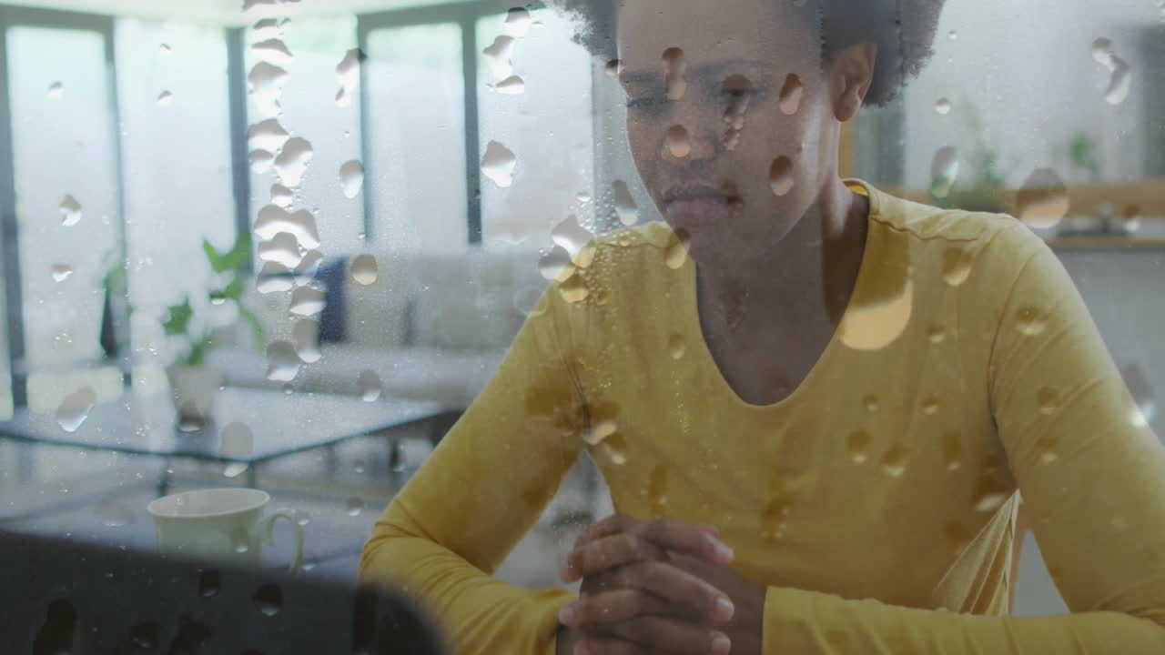 Animation of water droplets over unhappy african american woman sitting at home with head in hands
