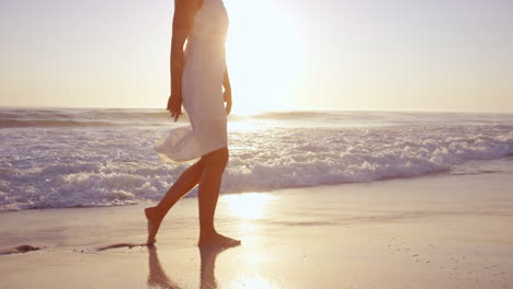 beautiful-woman-lifting-white-dress-walking-along-shore-line-on--beach-at-sunset-in-slow-motion-RED-DRAGON
