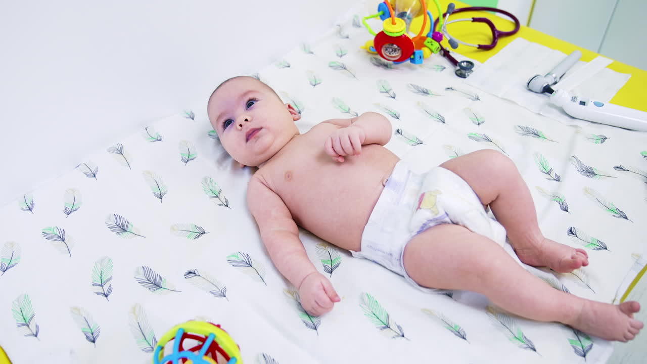 Naked baby in diaper only lying on white sheet. Medical devices are next to the baby. Bright toys around the child.