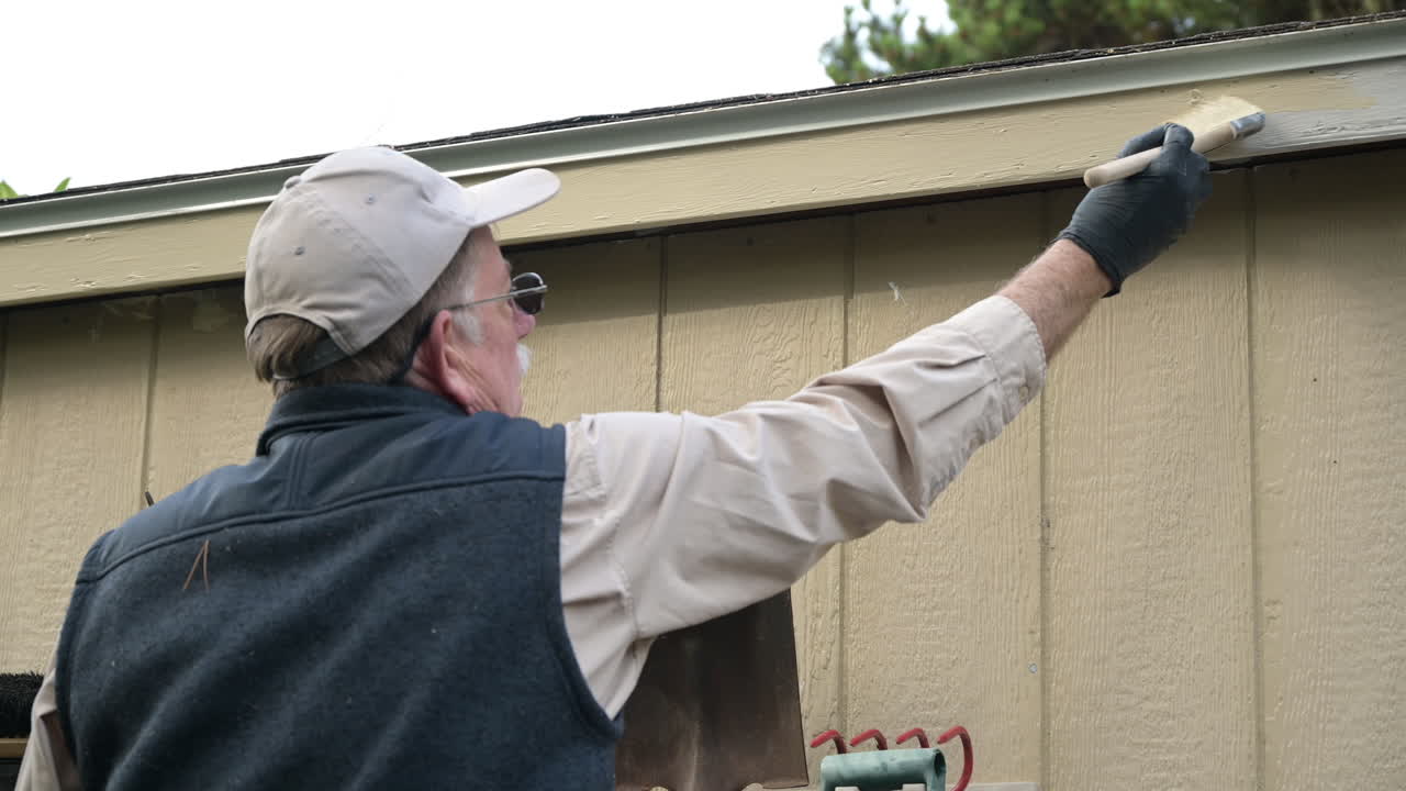 Elderly senior man paints trim on shed with paint brush