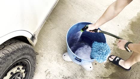 Video-Largo-De-Un-Joven-Blanco-Llenando-Un-Balde-Azul-Con-Agua-Y-Jabón-Preparándose-Para-Lavar-Un-Auto-Blanco-Sucio-Y-Fangoso