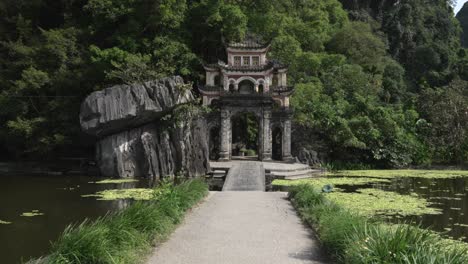 bich dong pagoda stone gate overlooks a lotus pond and limestone cliffs in ninh binh, vietnam; wind stirs foliage as a footbridge frames entrance, wide static handheld shot