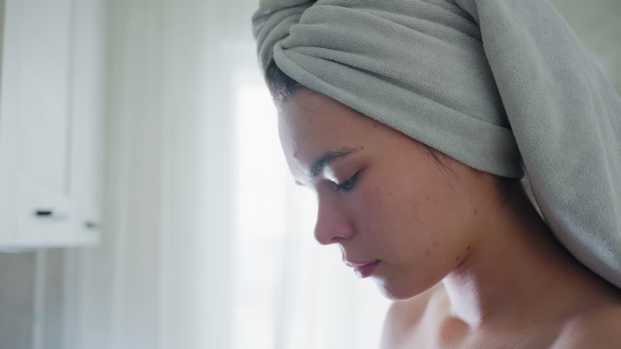 Close view of woman's face showing doted spots, focused expression, and towel-wrapped head, showcasing skincare and beauty routines, natural lighting highlights calm, relaxed atmosphere