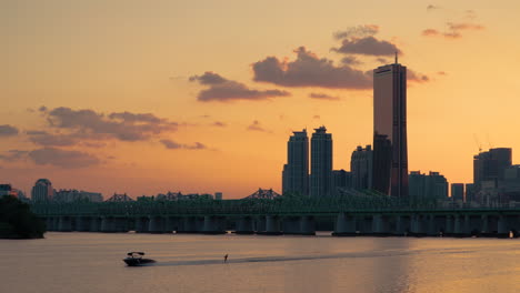 63 building in yeouido during sunset, train crosses on railway bridge, silhouetted man water skiing on han river when sunlight reflects in water