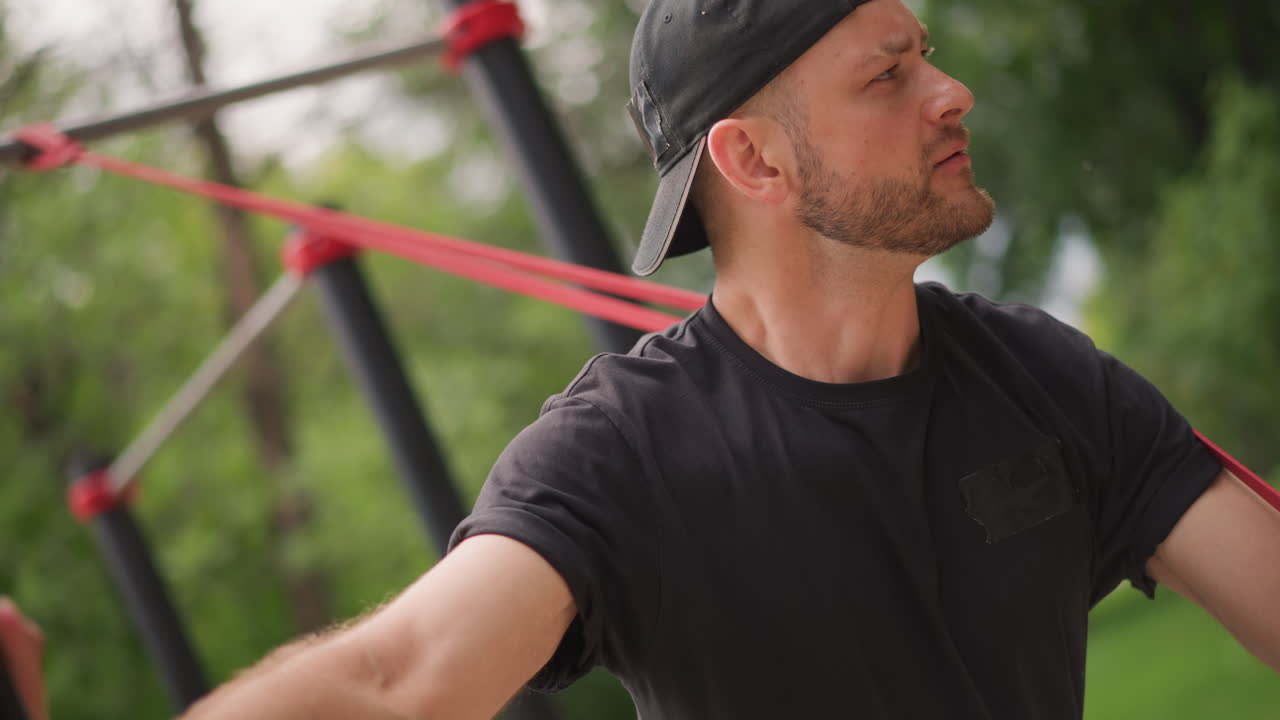 Man Performing Lateral Band Pulls For Upper Body In Park, Focused Athlete Extends Arms Wide Against Resistance, Controlled Tempo, Metal Posts And Leafy Background Create Urban Training Mood