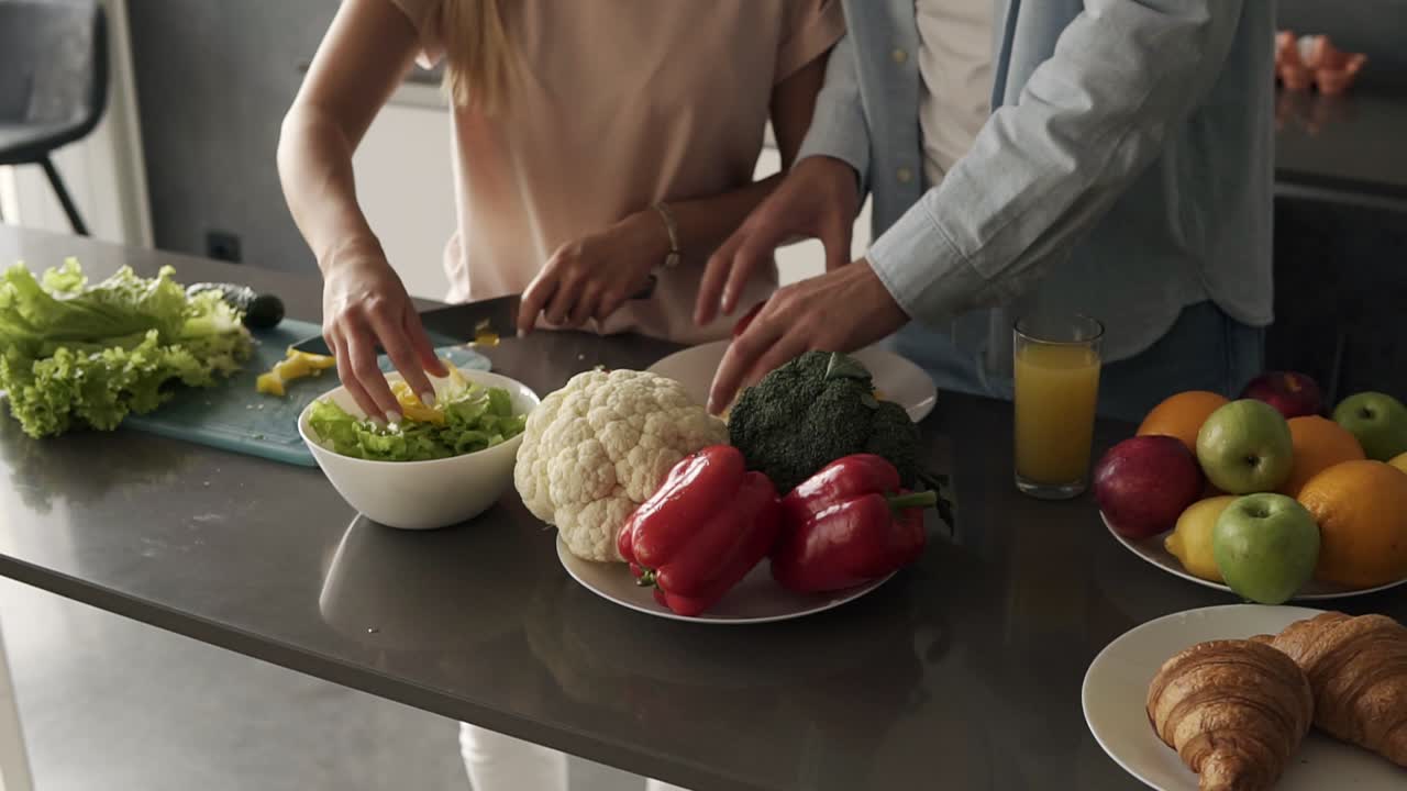 Unrecognizable young couple's hands cooking together a healty meal. Plenty of various colorful vegetables on the kitchen counter. Woman cutting salad on the cutting board, man breaking pepper slices on plate. Close up
