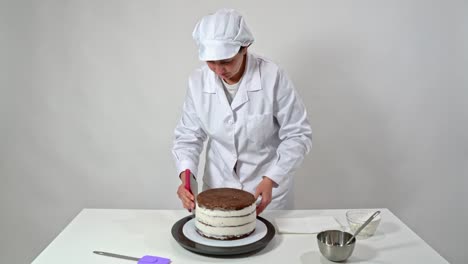 baker removes the excess cream from the sides of a chocolate cake by rotating the stand and using a spatula.