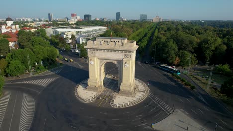 cinematic rotating view of the arch of triumph with the house of the free press in the distance, surrounded by lush green vegetation and tall office buildings, bucharest romania