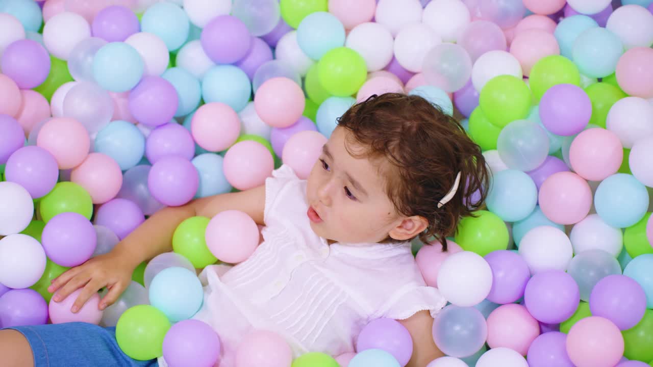 A 5-year-old girl rests and looks up, surrounded by colorful balls in an indoor ball pit