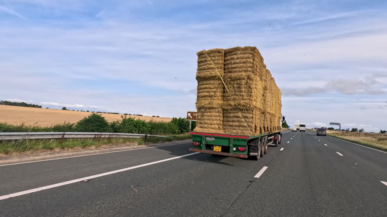 A large flatbed truck loaded with stacked hay bales travels along a multi-lane motorway in rural West Yorkshire under bright daylight, captured from a moving vehicle