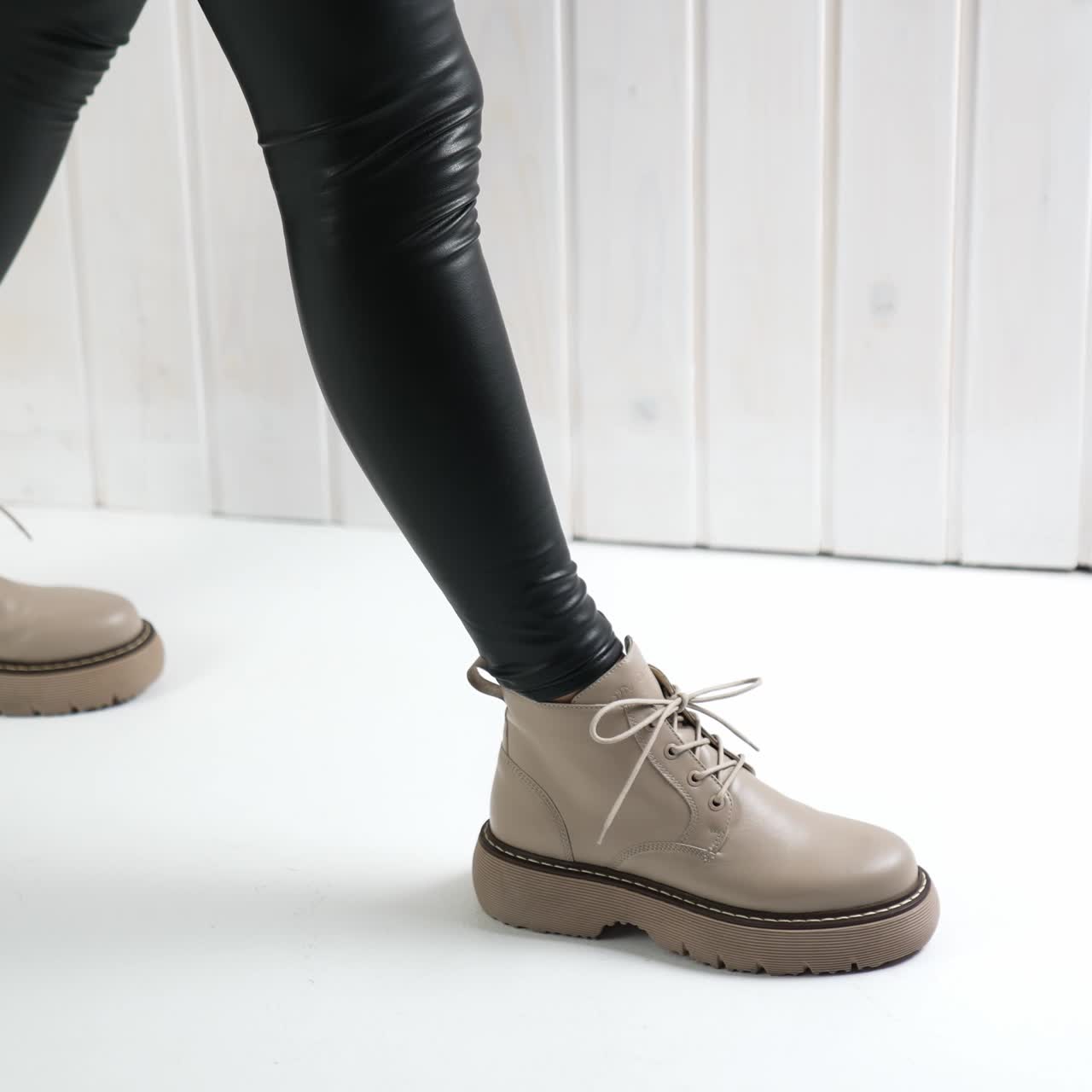 Lady in black leather leggings kneels to tie the lace on her stylish beige boots. Modern fashionable footwear show