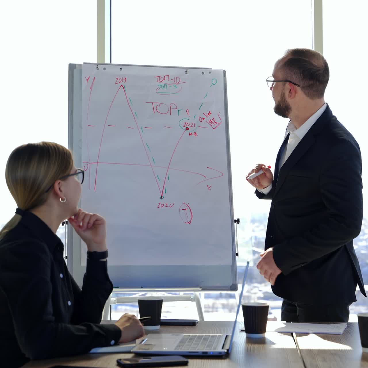 Smiling businessman standing near board and talking. Man leader in suit explaining company development strategy to colleagues showing on painted graphics