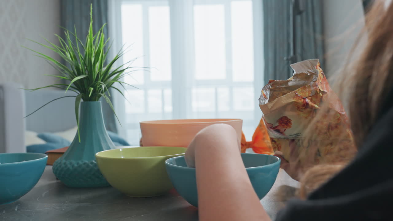 Close up view of person hand returning caramel popcorn to packet on dark table with colorful bowls on side and blurred person passing by in bright indoor setting, snack preparation moment
