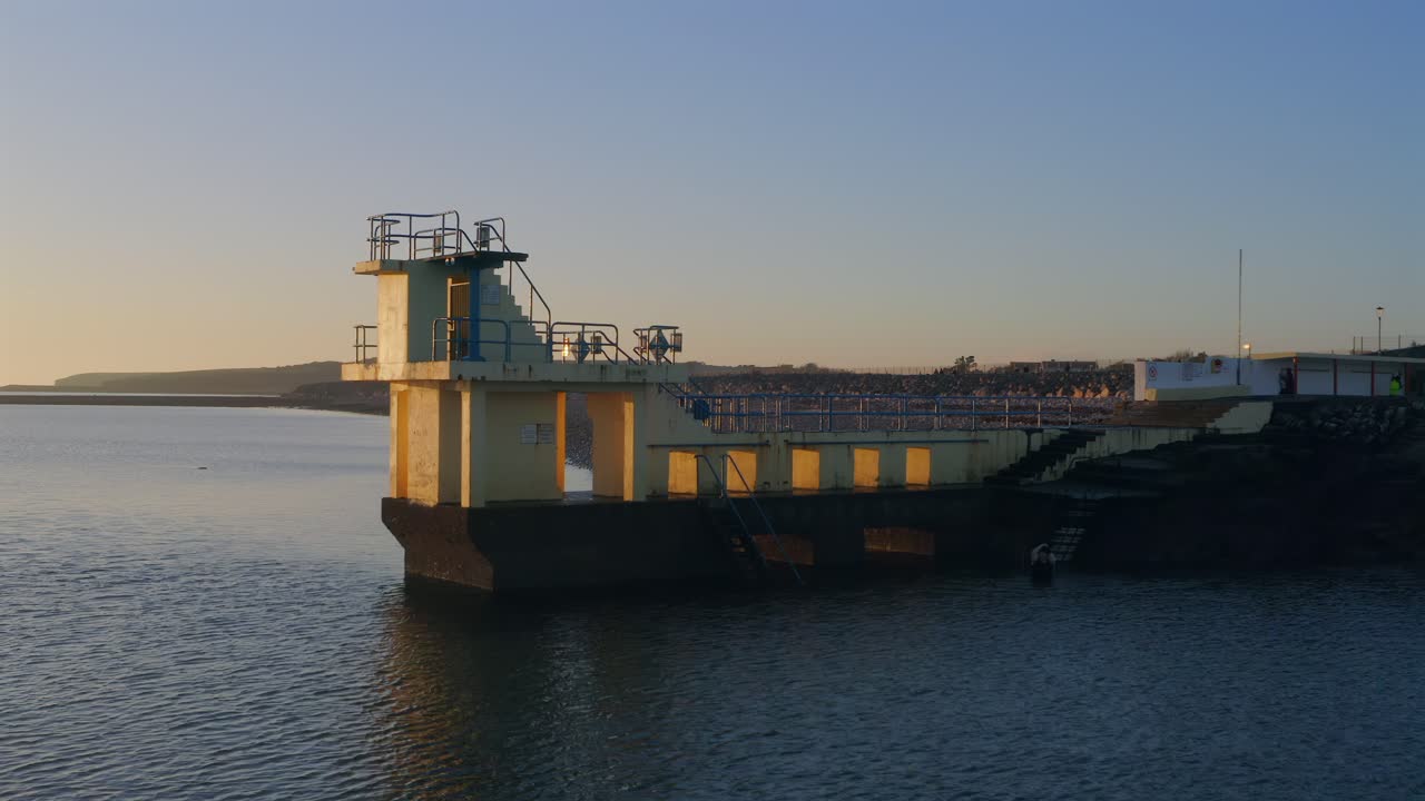 Aerial orbit of Blackrock diving board at sunset with swimmer entering the water.