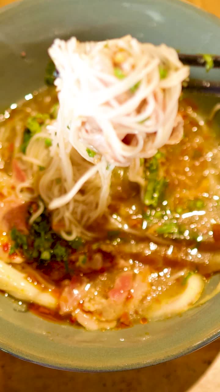温かい照明の環境で、箸が風味豊かなベトナムのスープから麺を持ち上げ、新鮮な食材と鮮やかな色彩が際立っています。