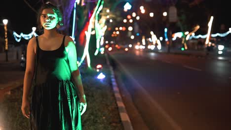 brunette woman in a black dress walking towards the camera in a street scene full of cars and colored lights