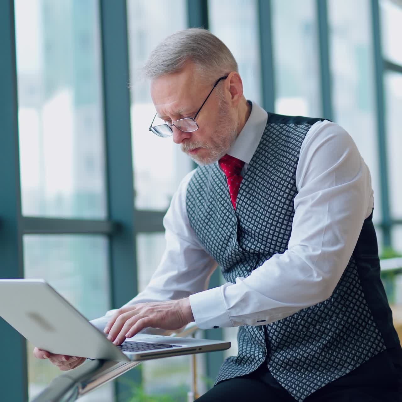 Nervous mature man working on a laptop. Serious senior businessman in eyeglasses standing near the window and looking at his laptop in his office.