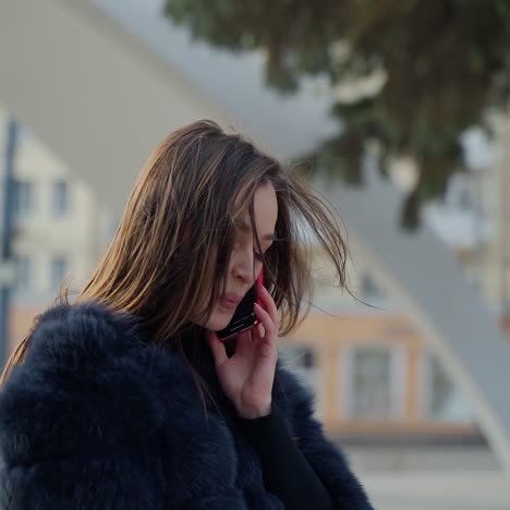 stylish girl with a phone outdoors. young brunette woman in fur coat walking and talking the mobile phone on the city background. slow motion.