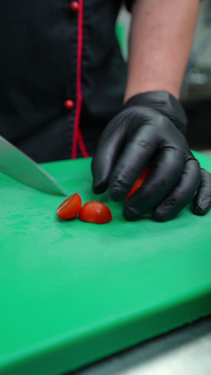 Vertical footage Professional chef wearing black gloves skillfully slicing fresh cherry tomatoes in half on a green cutting board, preparing ingredients for a delicious culinary creation