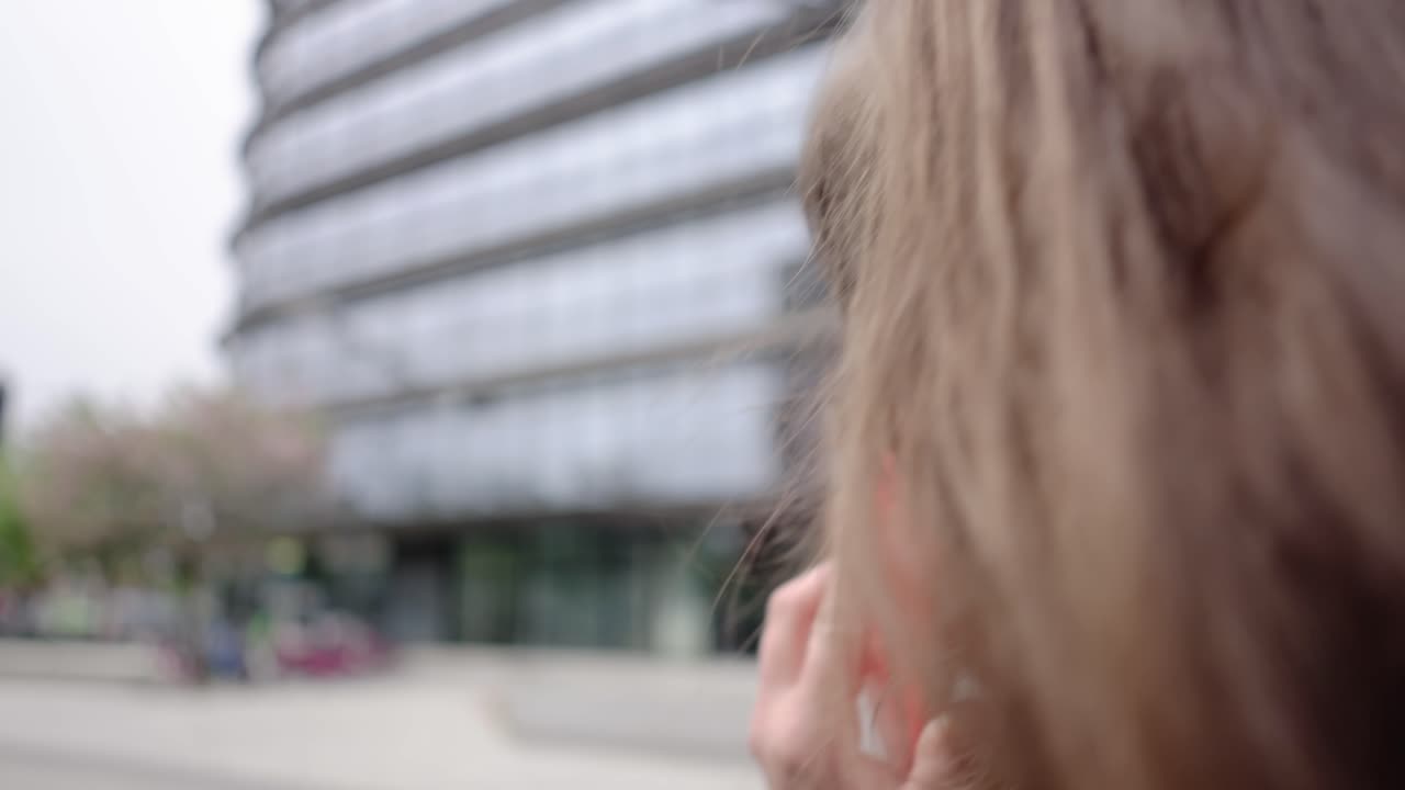 Close-up of a woman's head walking past a modern glass building, holding a red mobile phone to her ear