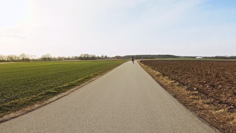 At-a-beautiful-day,-a-sportive-girl-is-inline-skating-along-a-rural-road-next-to-wide-fields-while-the-sun-in-shining,-filmed-from-behind