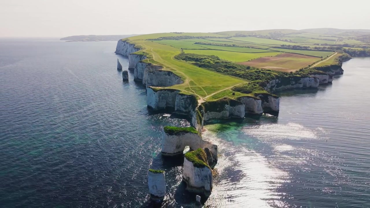 Ilmakuvaa dronella Old Harry Rocksista Dorsetin rannikolla Englannissa, esittäen luonnollisen merikaaren, valkoisia liitukallioita, meripilareita, vihreitä kallionhuippupolkuja, viljelysmaata ja tyyntä sinistä merta.