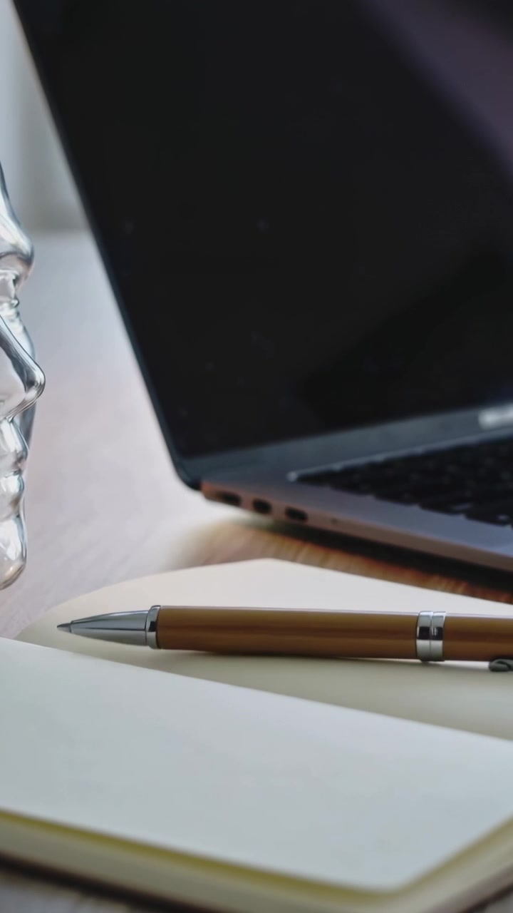 Video verticale: La telecamera scorre rivelando una penna marrone appoggiata su un quaderno aperto sulla scrivania, con un laptop.