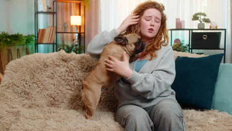 caucasian woman with headache sits on home sofa while pug dog cuddles face and shows empathy gently