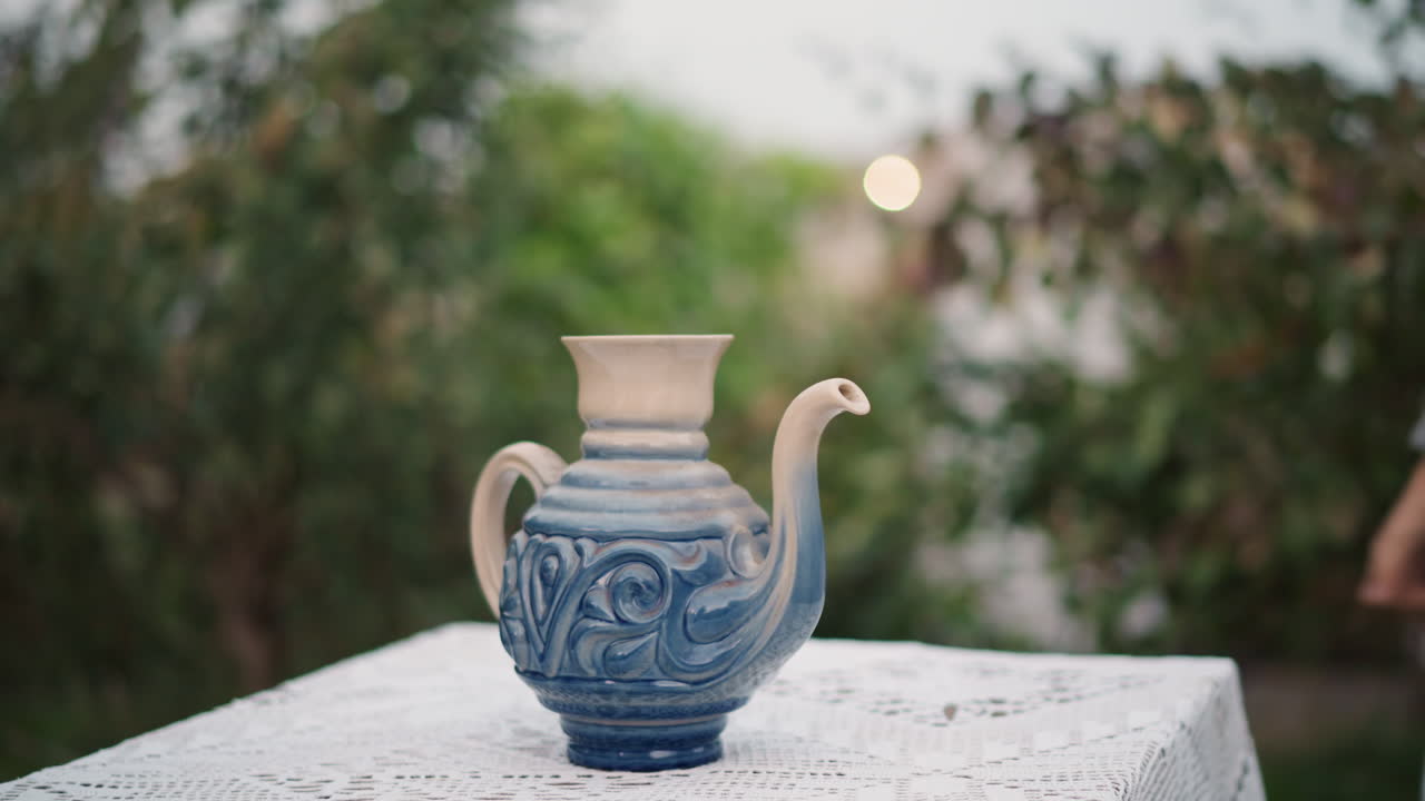 Hands placing ceramic blue jug on lace covered table outdoors person in gray knit sweater sets ornate indigo glazed teapot with decorative swirls, soft morning light, bokeh foliage background, careful
