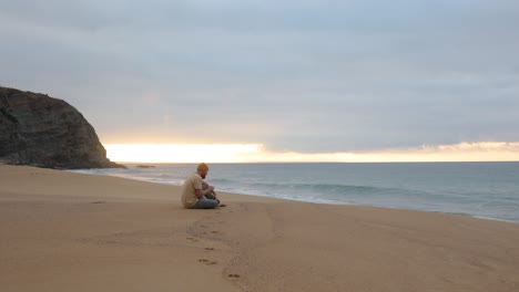 un hombre sentado y bebiendo café matutino mientras observa el amanecer en una playa aislada en australia