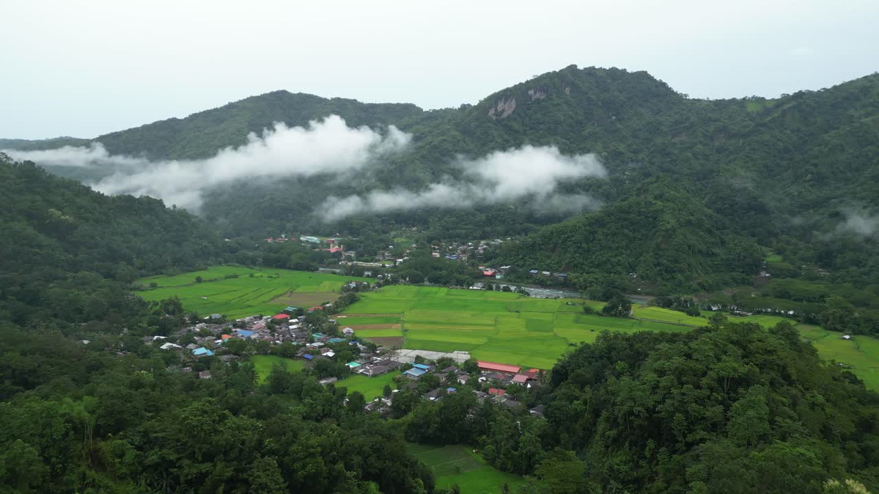 Uma vista aérea ampla captura um vale filipino exuberante com arrozais, casas de aldeia de telhado vermelho e colinas florestadas cobertas de névoa, misturando agricultura e natureza em harmonia cinematográfica