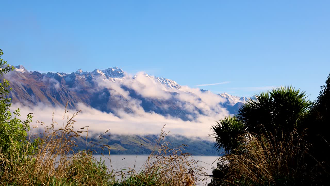 Snow-capped mountains and fog over Lake Wakatipu with clear blue skies create a tranquil scene in New Zealand
