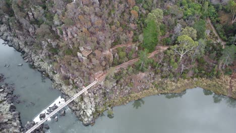 4K-Aerial-view-of-a-Gorge-with-footpaths,-a-suspension-bridge-and-a-small-waterfall-in-an-Australian-National-Park