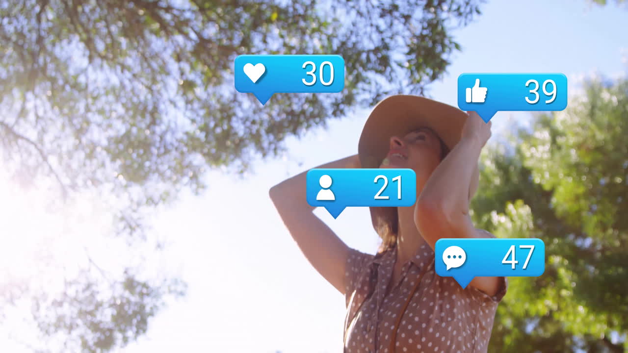 Woman adjusting straw hat in park, showcasing floating heart and thumbs-up icons for marketing