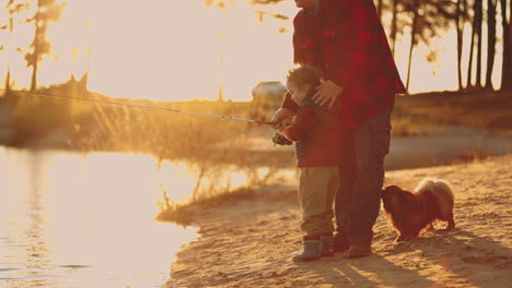 little boy father and dog are resting on shore of lake in sunset child is learning to catch fish