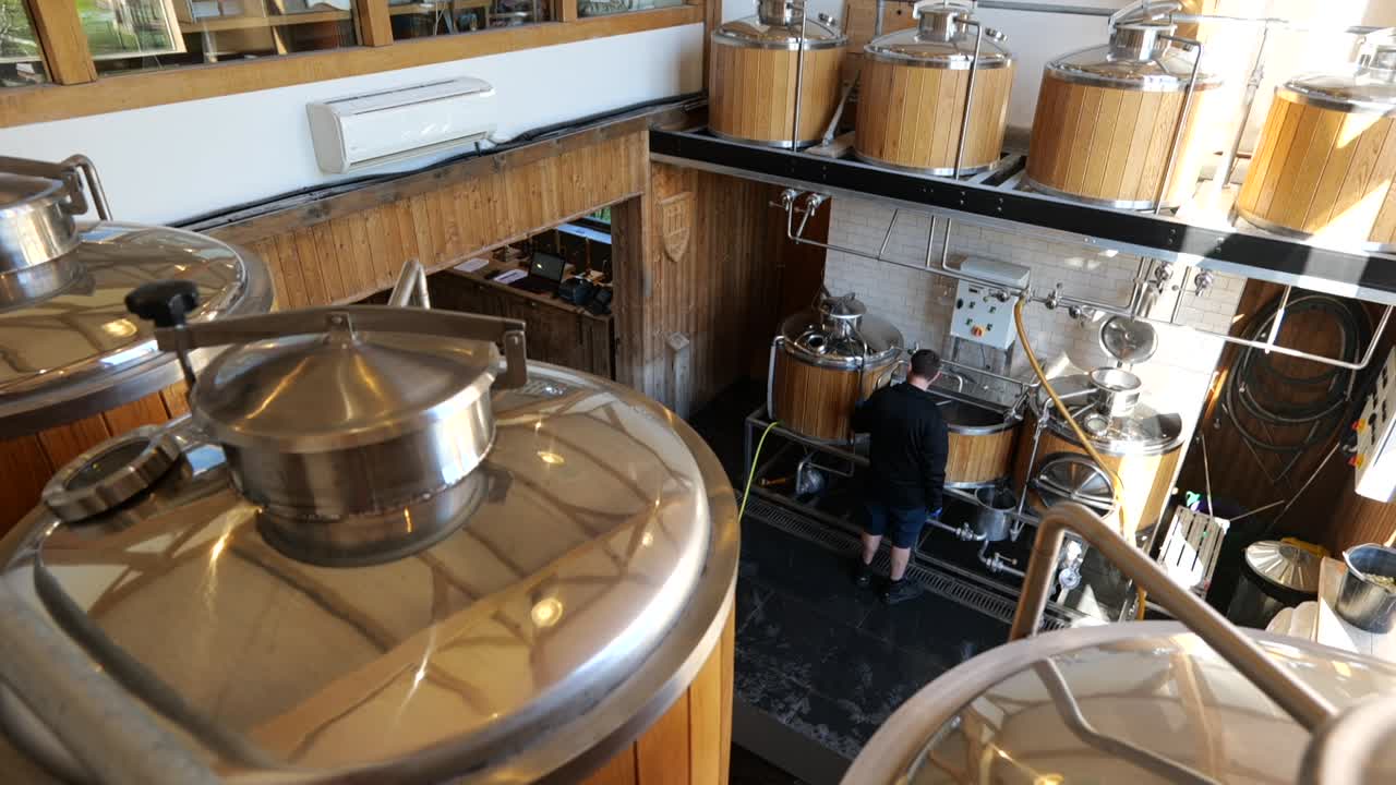 Movimento panorâmico para a esquerda em ângulo de três quartos, mostrando cervejeiro inspecionando tanques de cervejaria, interior com luz natural.