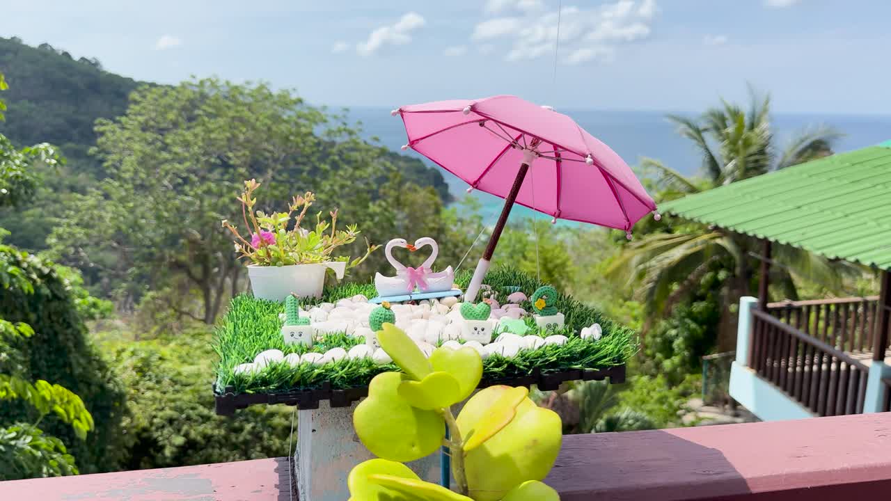 Vibrant swan toys and pink umbrella on a clifftop in Phuket, Thailand. Bright daylight highlights lush greenery and ocean views