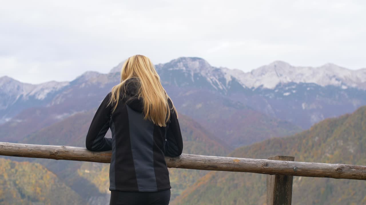 Blonde young woman leaning on wooden fence looking towards mountains
