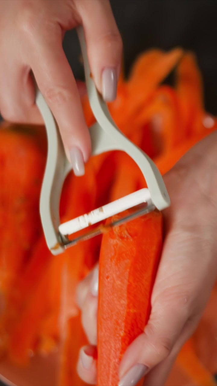 Woman hands use vegetable peeler to remove skins from bright orange carrots revealing fresh peeled pieces. close up view emphasizes texture of carrot skin and smooth peel against blurred