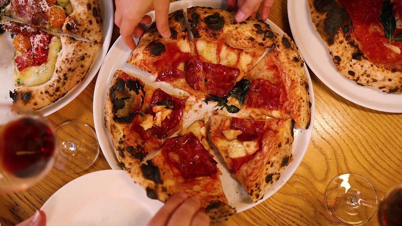 Scatto dall'alto di persone che prendono fette di pizza fresca al salame piccante da un tavolo di legno di un ristorante.