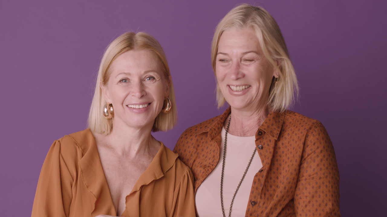 dos mujeres rubias maduras con pantalones marrón abrazándose y sonriendo con fondo morado