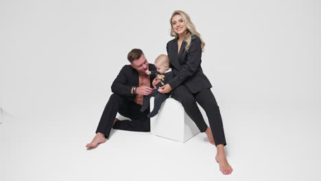 beautiful smiling blonde lady in black suit sitting on white cube. little kid sits beside supported by dad. studio footage of happy family. white backdrop.