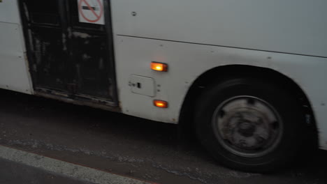 una vista cercana de un coche que pasa por una carretera húmeda con reflejos, mientras que un autobús blanco con una insignia de no fumar en la puerta, se detiene cuando su puerta se abre