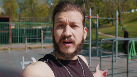 a man with a beard is taking a selfie at the outdoor gym. he is wearing a black tank top and a chain. he is showing off his muscles and looks like he is ready to work out.