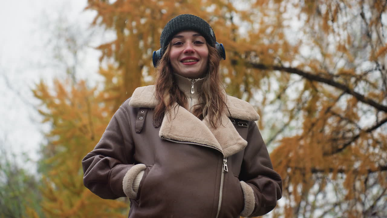 Smiling female wearing black knit cap and brown shearling jacket with hands in her pockets outdoors during cool autumn day, enjoying the moment in nature